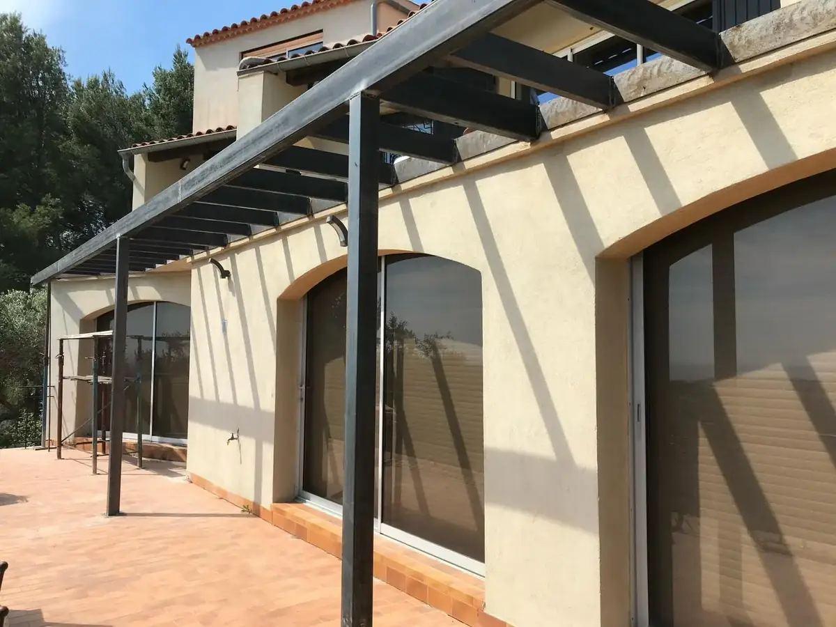 Extension de balcon d'une villa : ossature en acier noir sur poteaux, fixée en façade au-dessus d'une terrasse carrelée bordée de baies vitrées cintrées