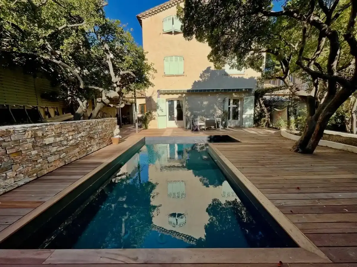 Piscine rectangulaire créée dans un jardin de Toulon, bordée d'une terrasse en bois et d'un mur habillé de pierre, devant une maison aux volets vert pâle