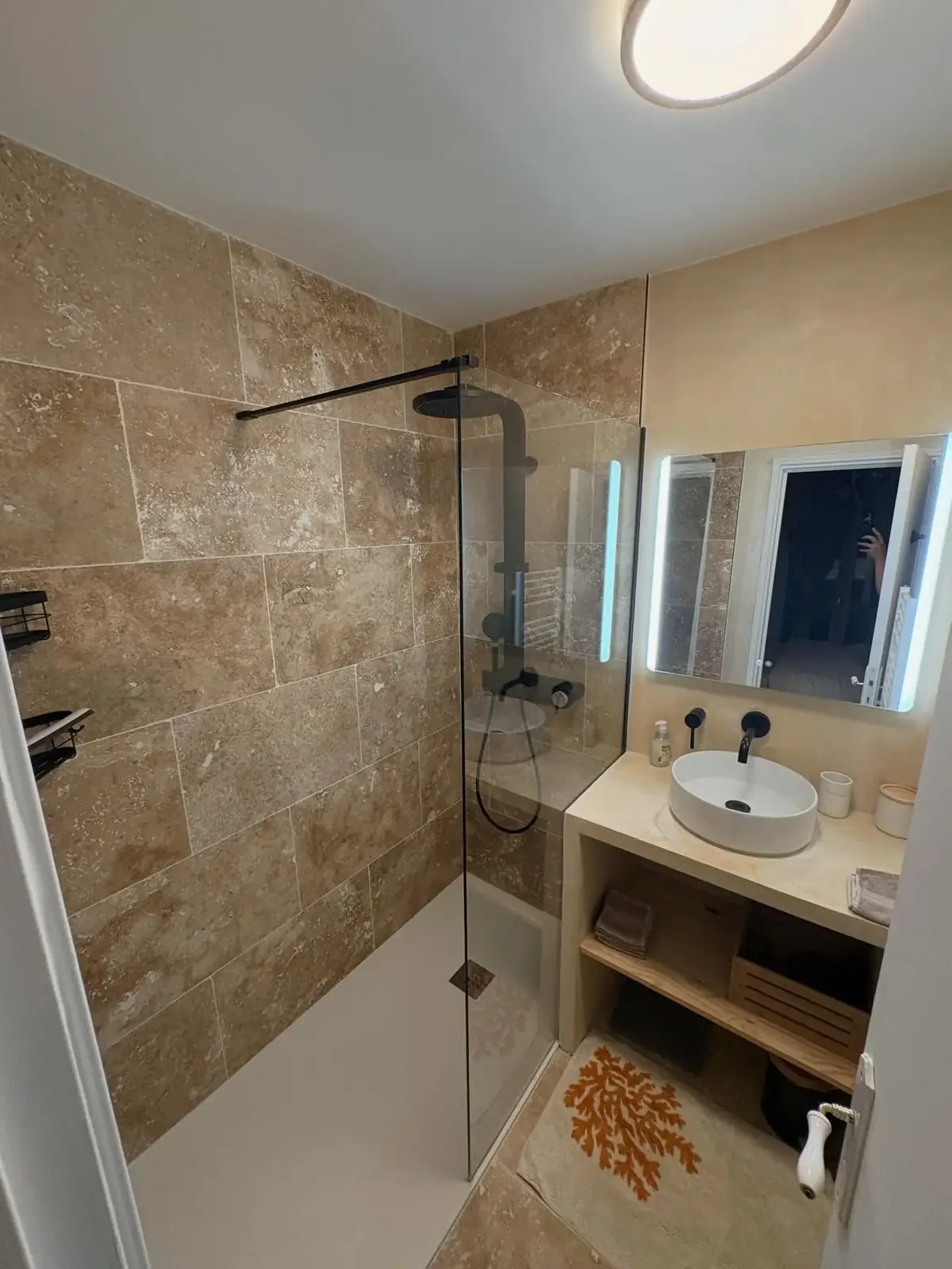 Salle de bain rénovée par L'Atelier du Golfe : murs en travertin, douche à paroi fixe, vasque sur meuble maçonné et miroir lumineux