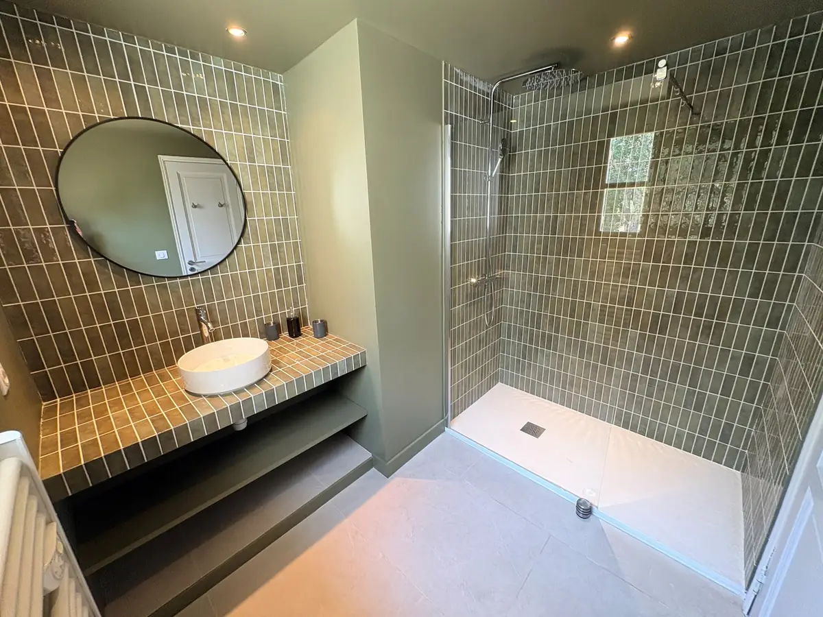 Salle de bain rénovée : zellige vert sur la douche ouverte et le plan vasque, paroi vitrée fixe, vasque à poser et miroir rond