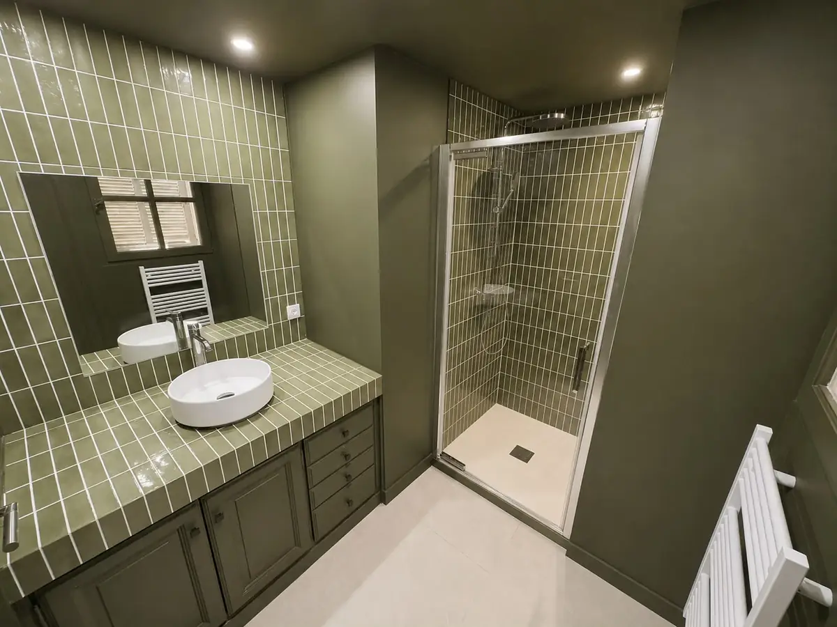 Salle de bain rénovée : zellige vert olive posé verticalement sur le mur et le plan vasque, vasque blanche à poser, douche à paroi vitrée et sol en carrelage clair