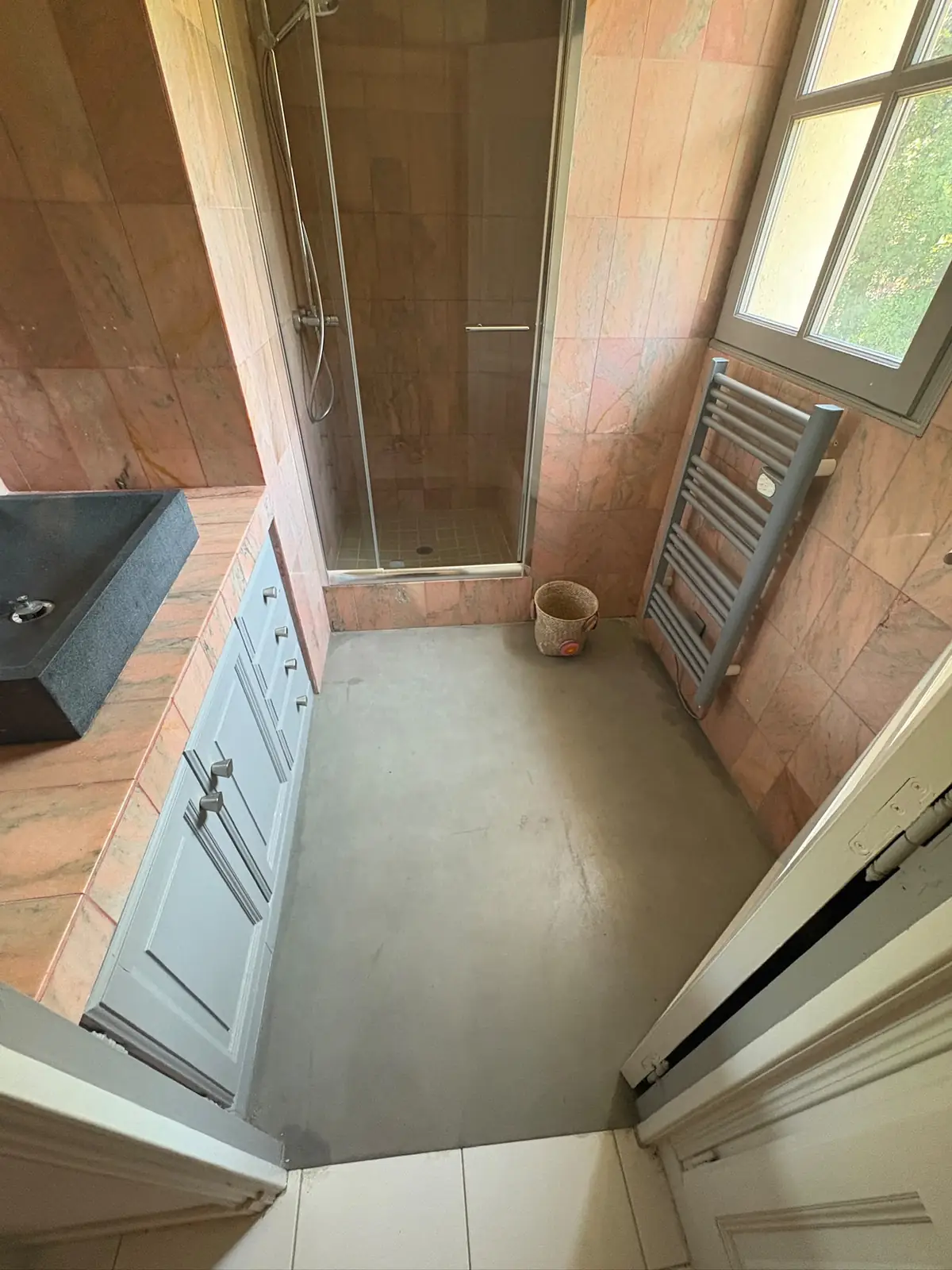 La salle de bain avant travaux : murs et plan vasque en marbre rose, cabine de douche et sol gris